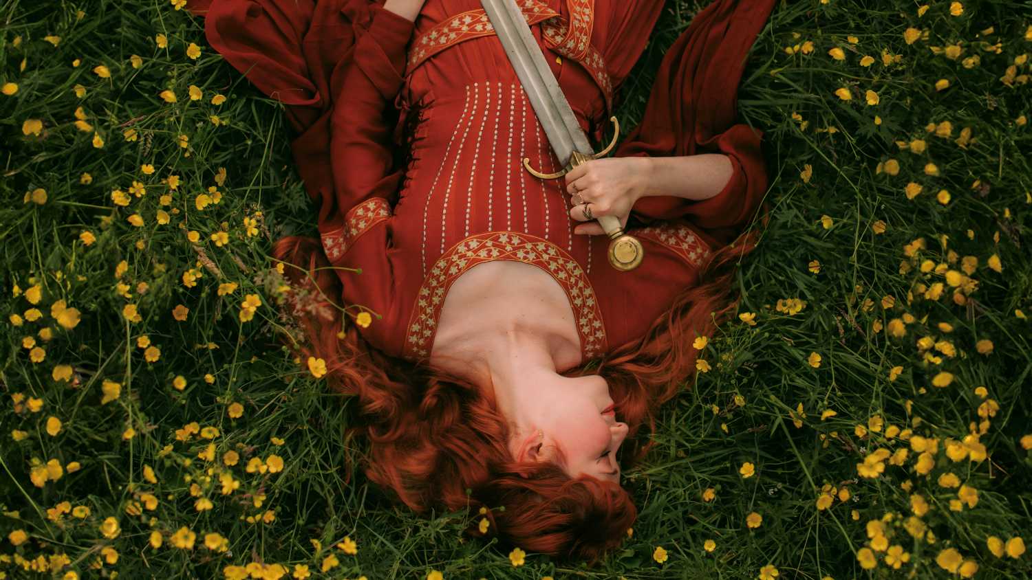 beautiful woman lying in bed of yellow flowers while holding a steel sword