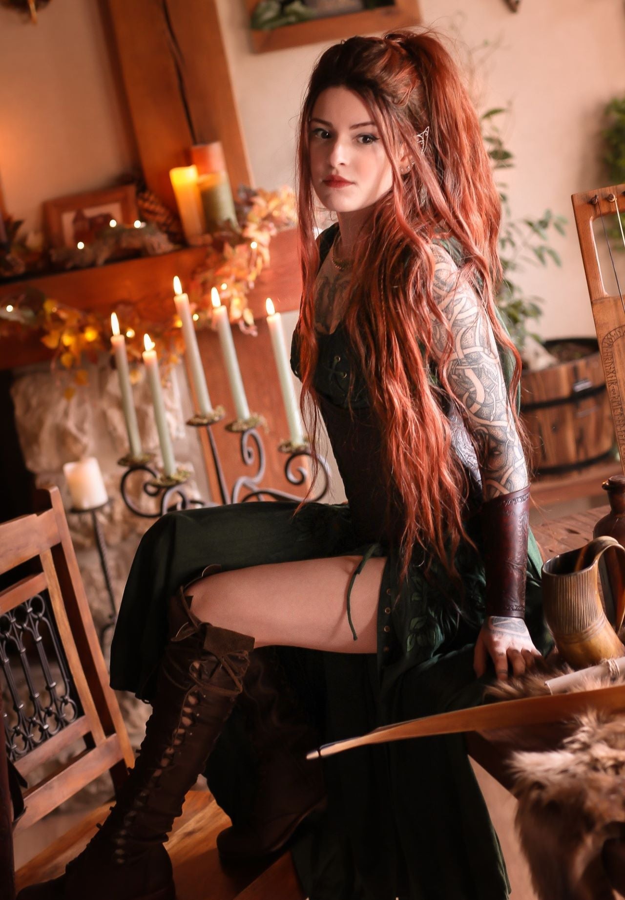 Red-haired woman in green medieval elf costume with full sleeve tattoos by candlelit fireplace